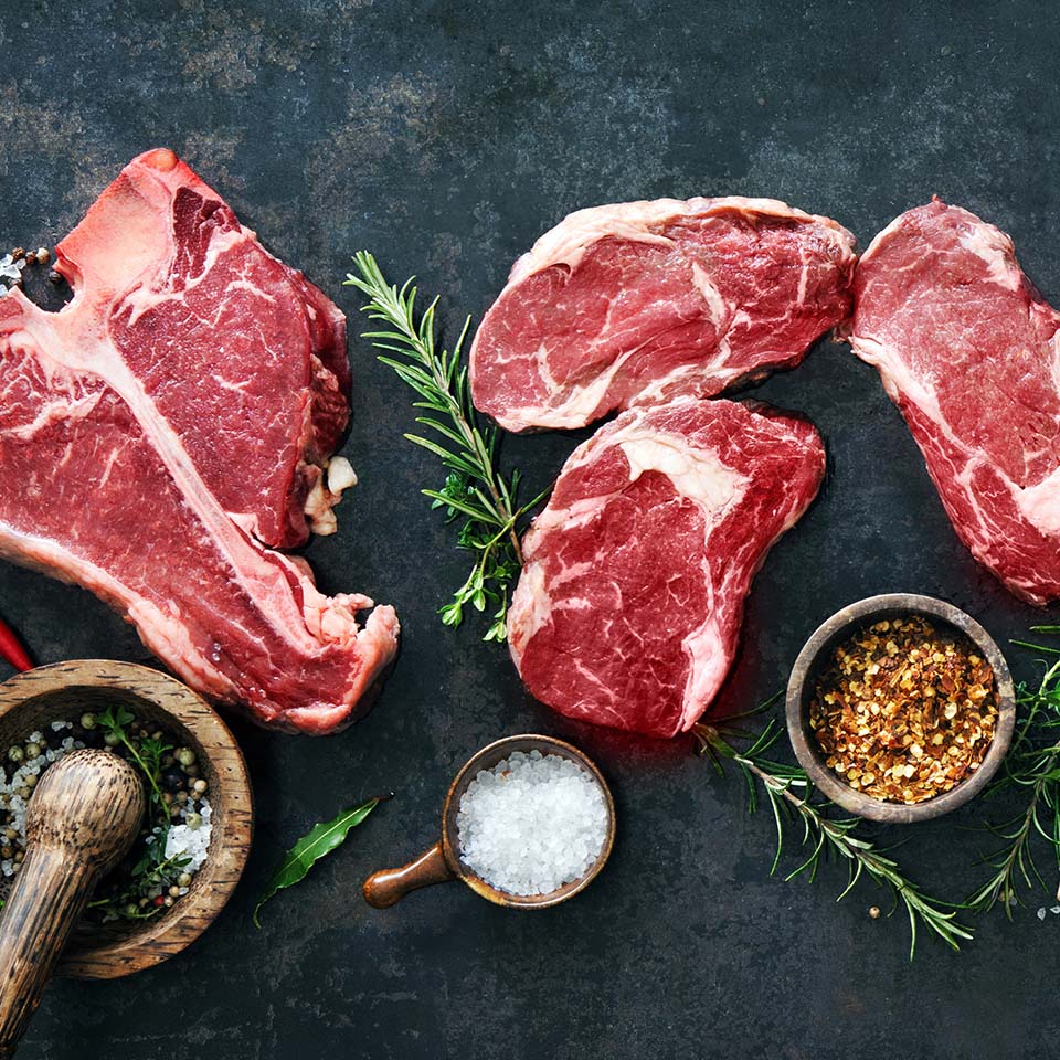 Variety of raw beef meat steaks for grilling with seasoning and utensils on dark rustic board